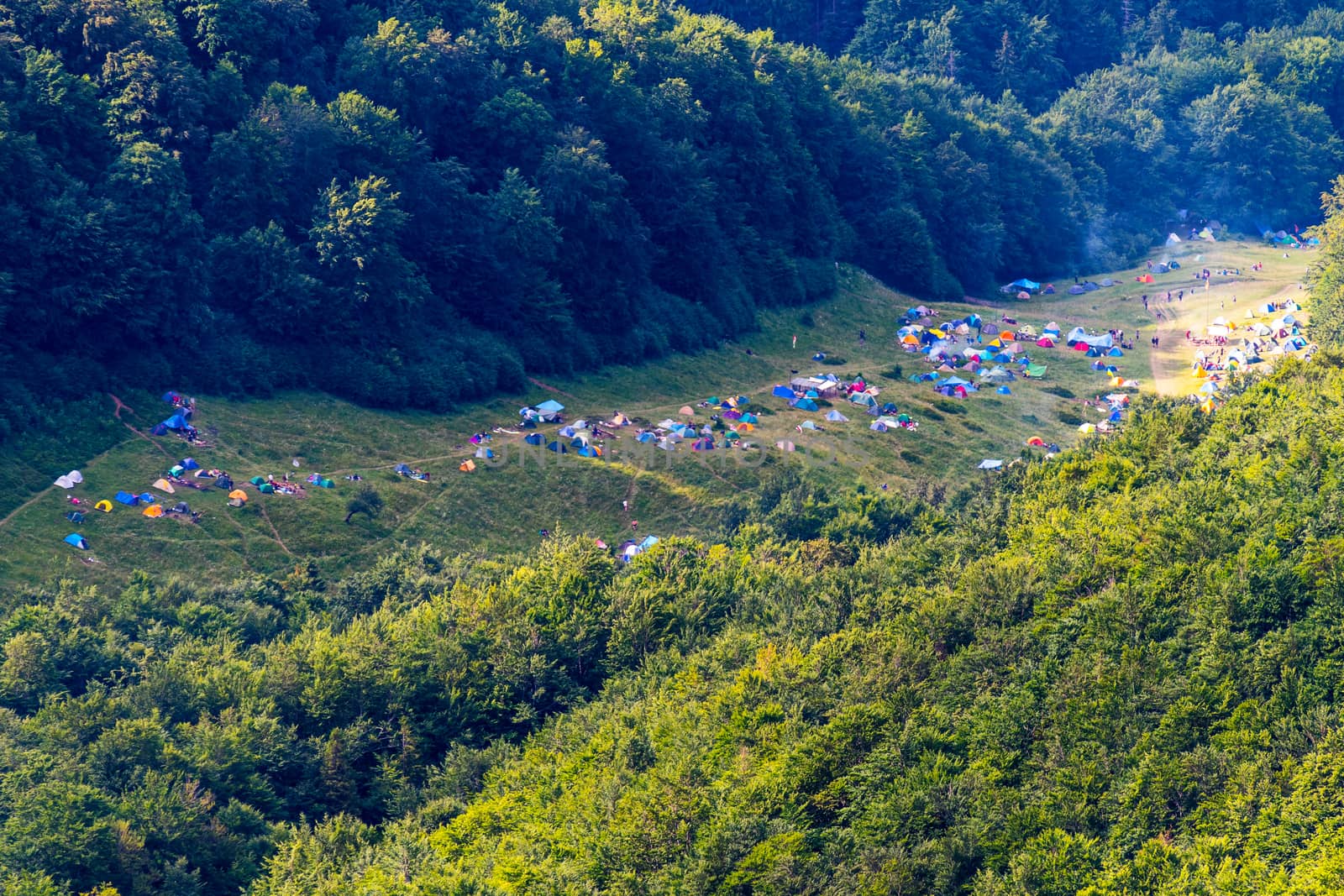 Multicolored spots on the green grass in the valley between the slopes of the overgrown trees is a tent camp located there. by Adamchuk