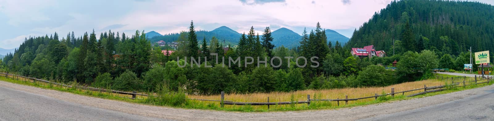 Quirky asphalt road with a wooden fence and a view of small houses and hotels amidst large green mountains by Adamchuk