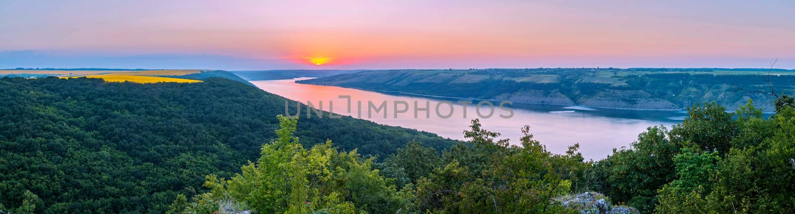 A magnificent panorama of a wide river flowing between the green beaches. With yellow squares of wheat crops and the sun on the horizon rolling down. by Adamchuk