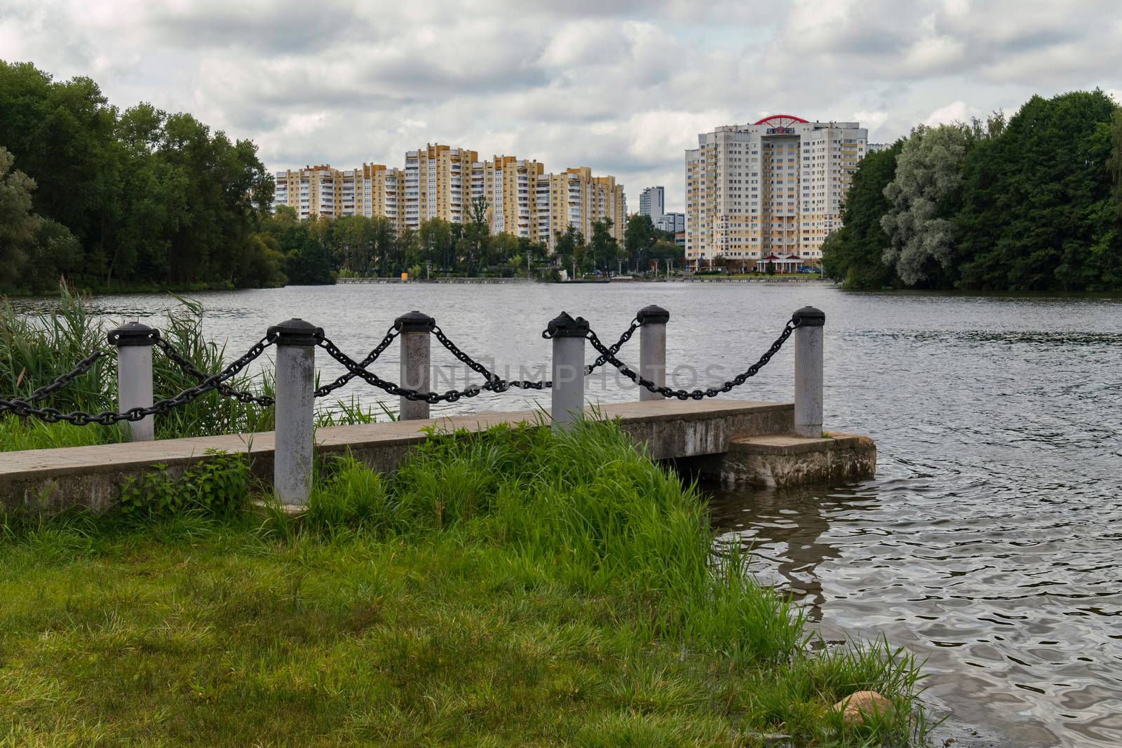 A small pier with chain fences on a city water body in the foreground and a high-rise building in the back