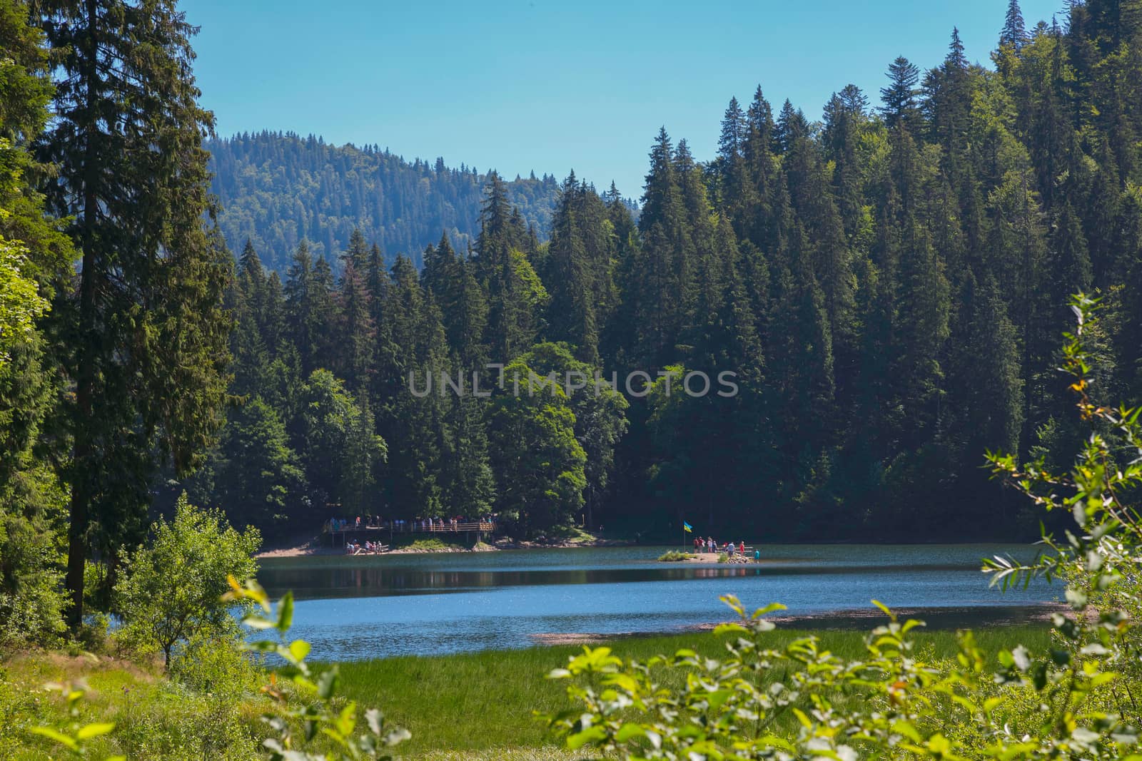 Tourists on a small island with a flag fluttering in the midst of a mountain lake surrounded by a tall and dense forest.