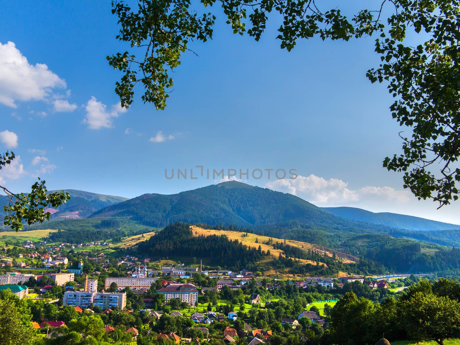 A beautiful landscape of the city on a summer day lying in a mountain valley against the background of yellow slopes of wheat and a peak overgrown with green forest.