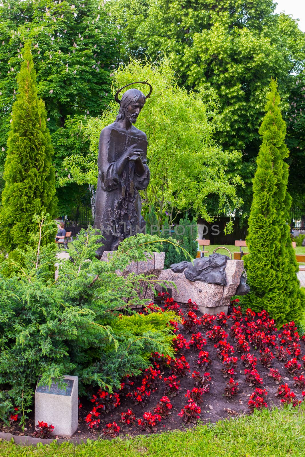 Sculpture of a praying saint with closed eyes standing near a flower bed with beautiful flowers against the background of green trees.