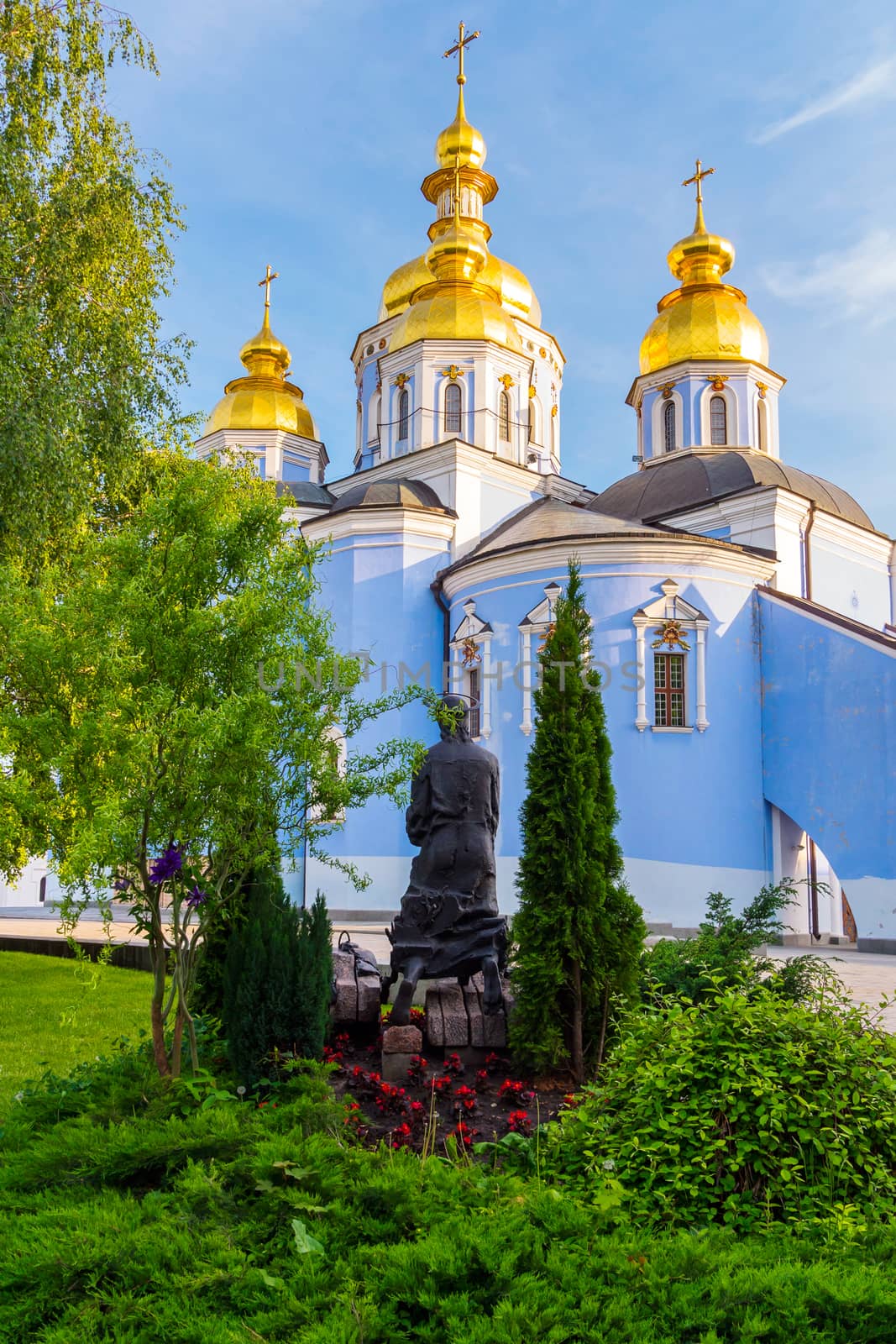 A small sculpture standing near a beautiful church with domes shining gold in the sun and crosses on them.