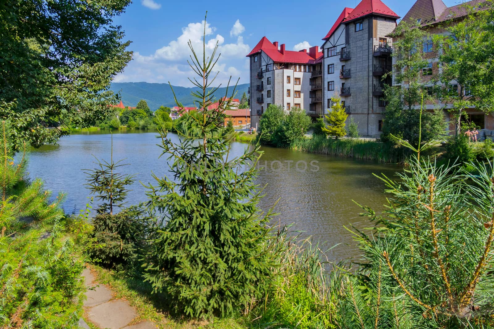 A multi-storey building with a red roof on the shore of a small lake with trees by Adamchuk