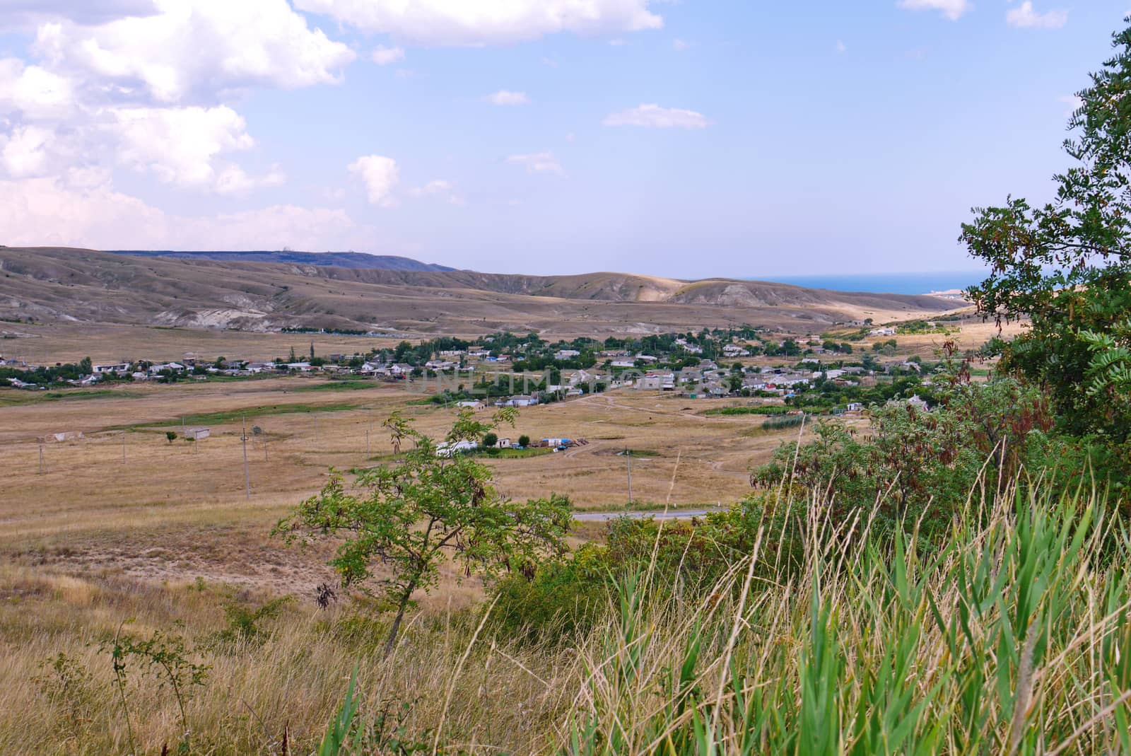 A small village in the lowland of a hill with dried grass around and a sea that is visible in the distance.