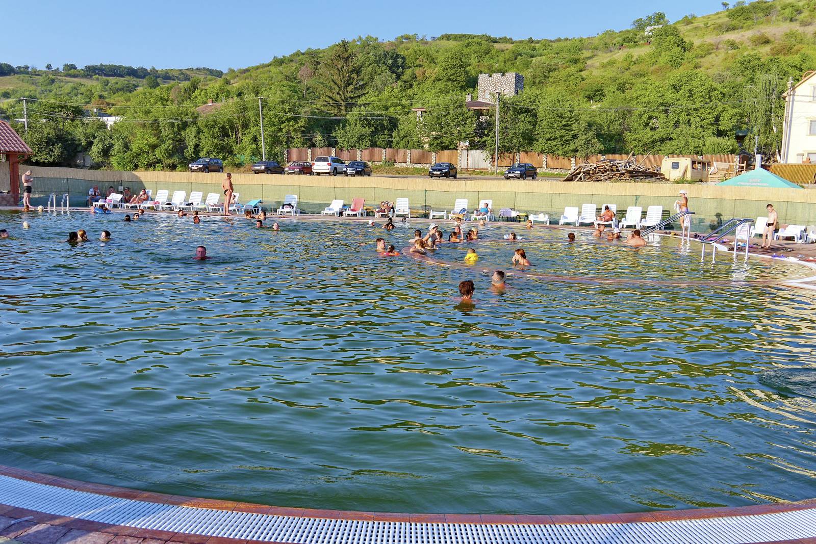 Tourists are swimming and refreshing in the pool on a hot summer day. Overlooking a slope covered with green trees and parking with cars.