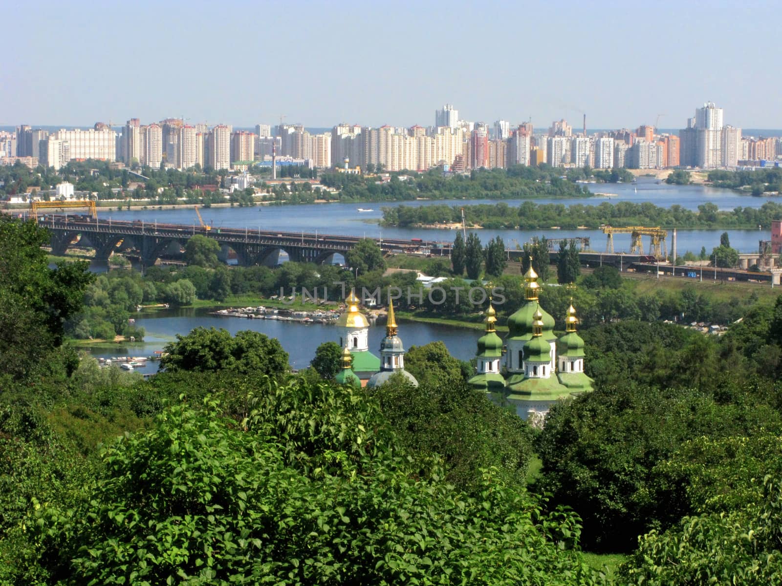 An excellent landscape with the tops of domes over green trees. A bridge going through a wide river and city blocks of high-rise buildings.