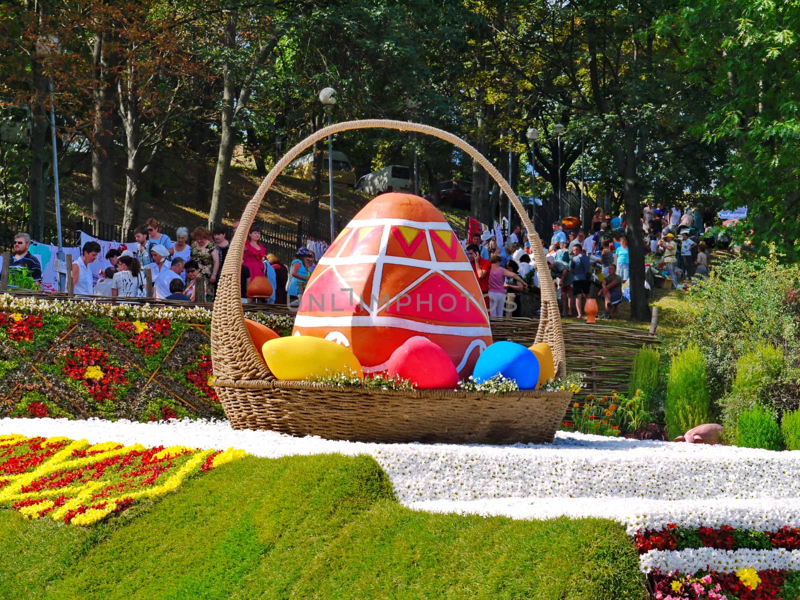 Exhibition is a summer sunny day in the park. A large wicker basket with a painted egg standing on a white veil of flowers and people looking at this beauty.