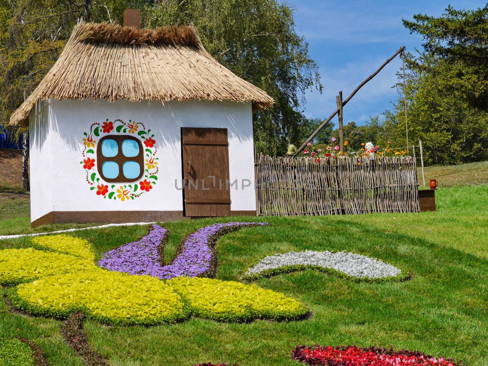 Authentic Ukrainian hut with a fence, a well and magnificent flower beds in the front yard on the background of the forest.
