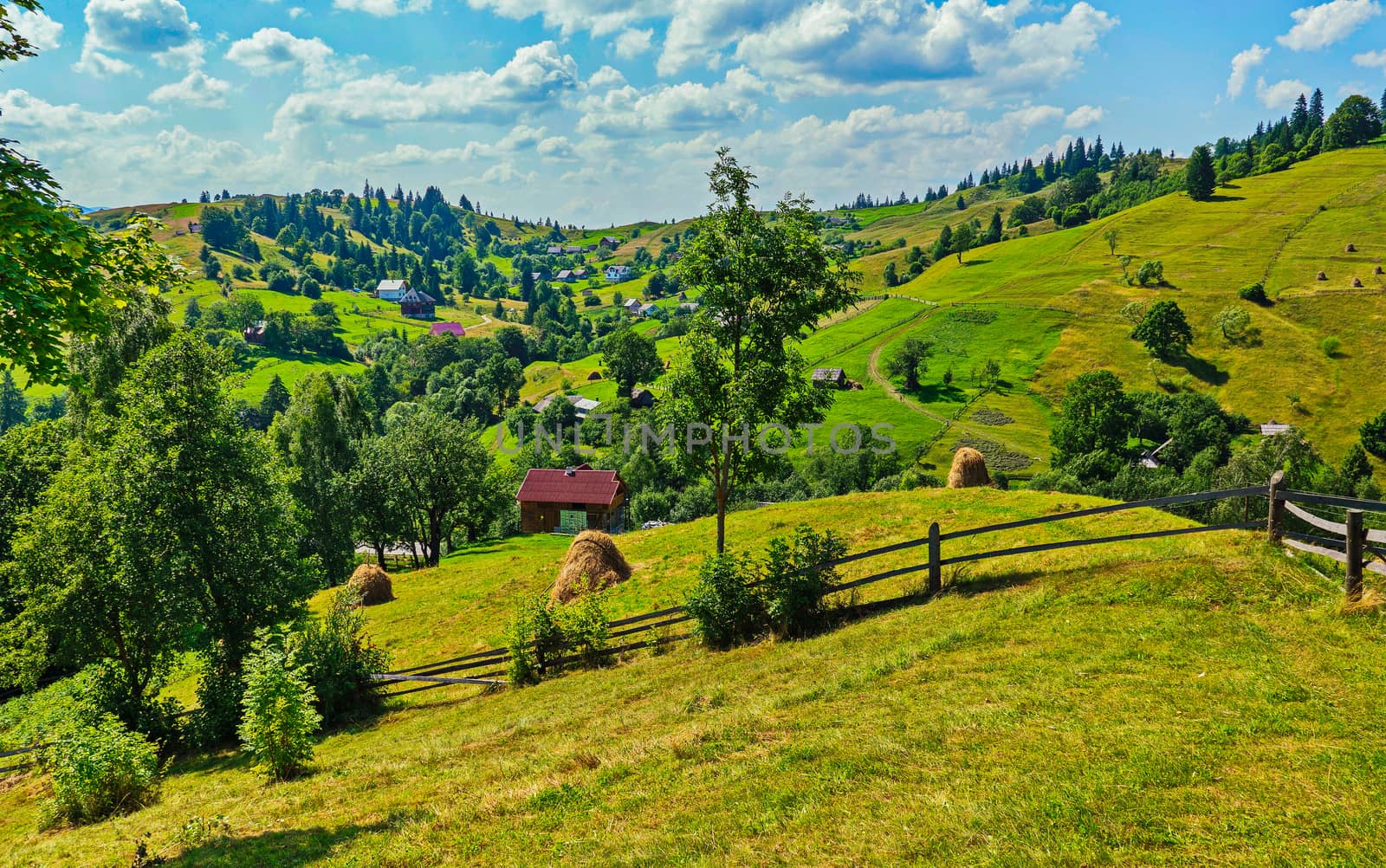 A picturesque plain with huts and haystacks against the background of green mountains and blue sky