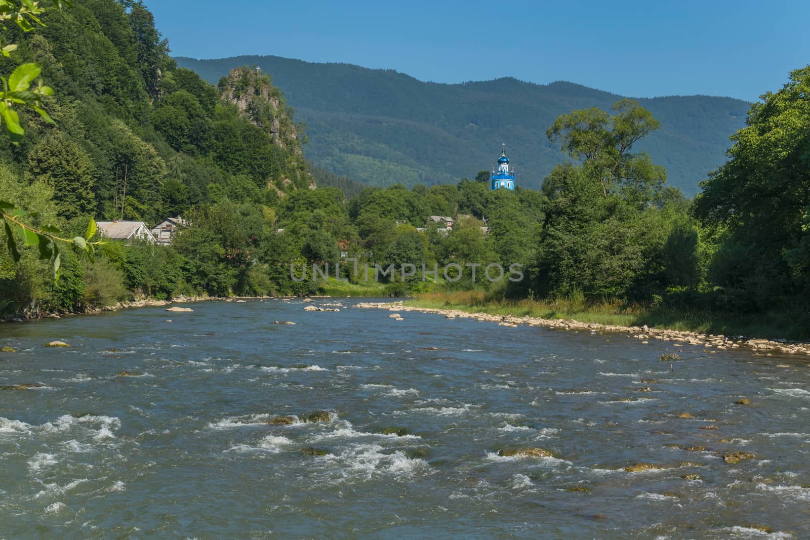 The swift mountain river against the background of the blue top of the church church, small houses and rocky mountains