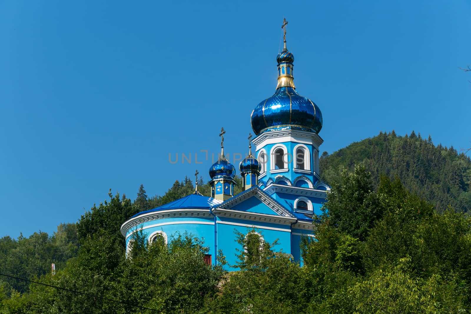 A beautiful church with a shining blue in the sun with a dome standing in the green thickets of trees against the background of a cloudless blue sky.