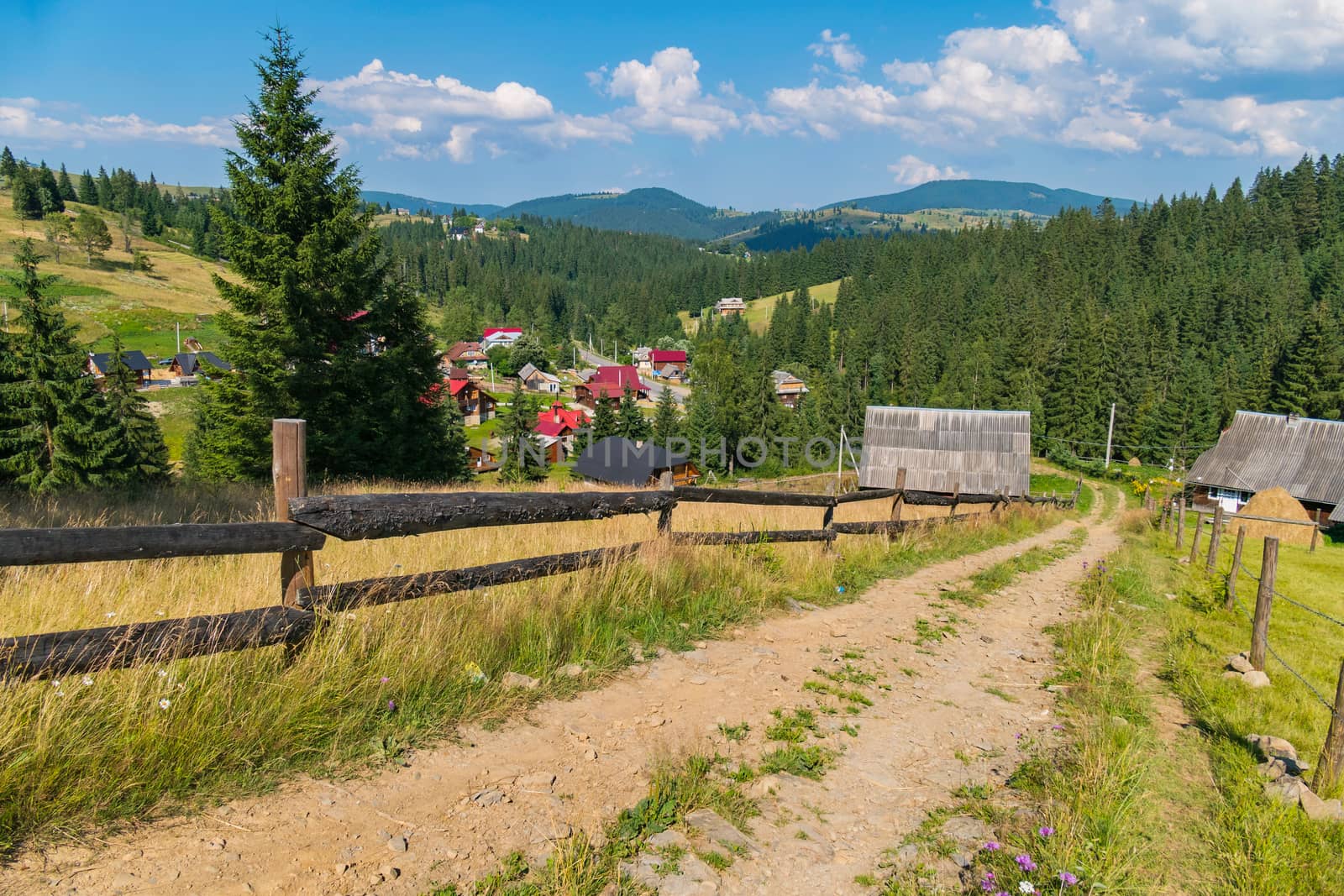 A sandy dirt road in the countryside running between the wooden hedges to houses in the distance.