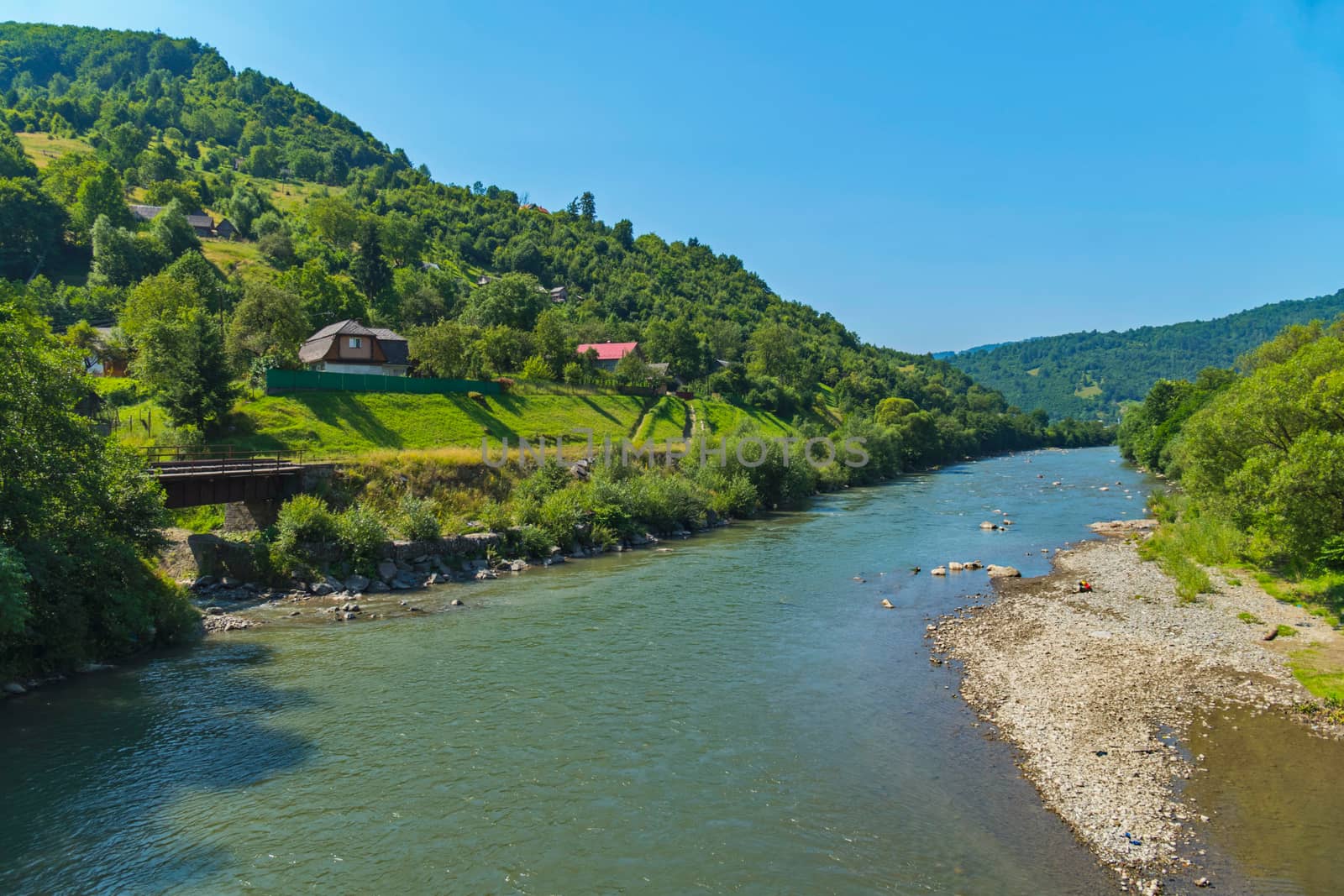 wide mountain river with tributaries and rocky shores near the village on the background of green hills
