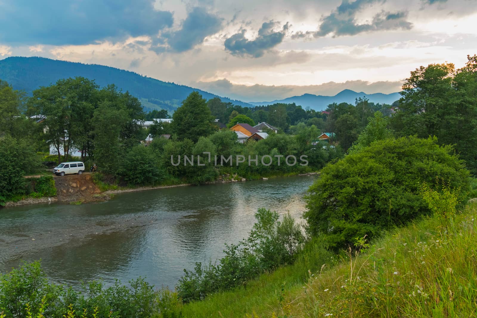 roofs of rural houses among green trees on the bank of the river with a white minibus parked next to it against the backdrop of distant peaks and a gray cloudy sky. place of rest and tourism