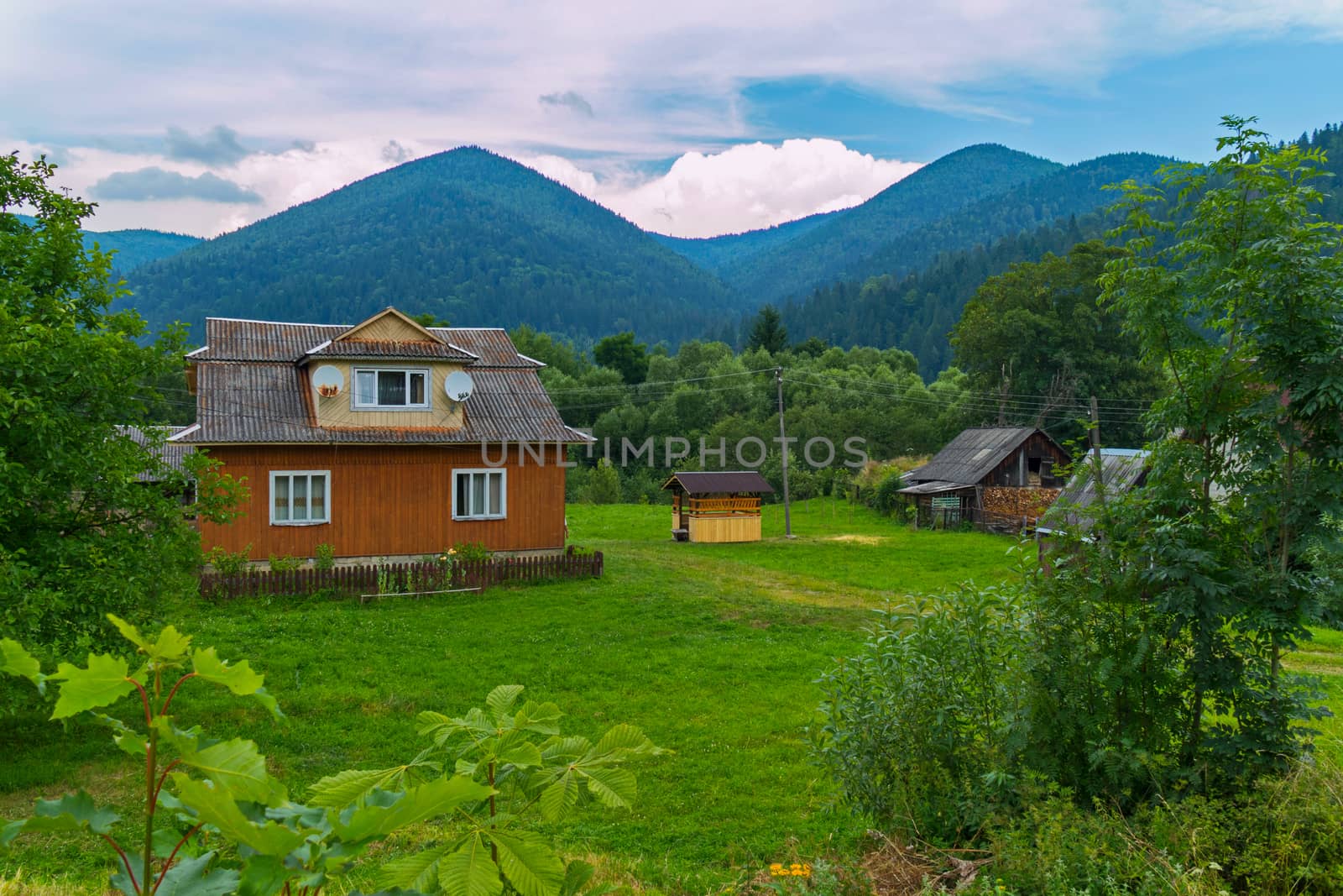A small rural house with a courtyard and an old well against the background of green mountains and white clouds
