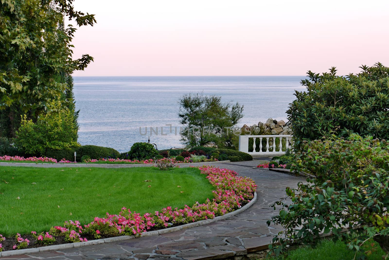 The path in the park is paved from a large variety of plates enveloping a green lawn with a flower bed on one side and with a beautiful view of the sea from the other.