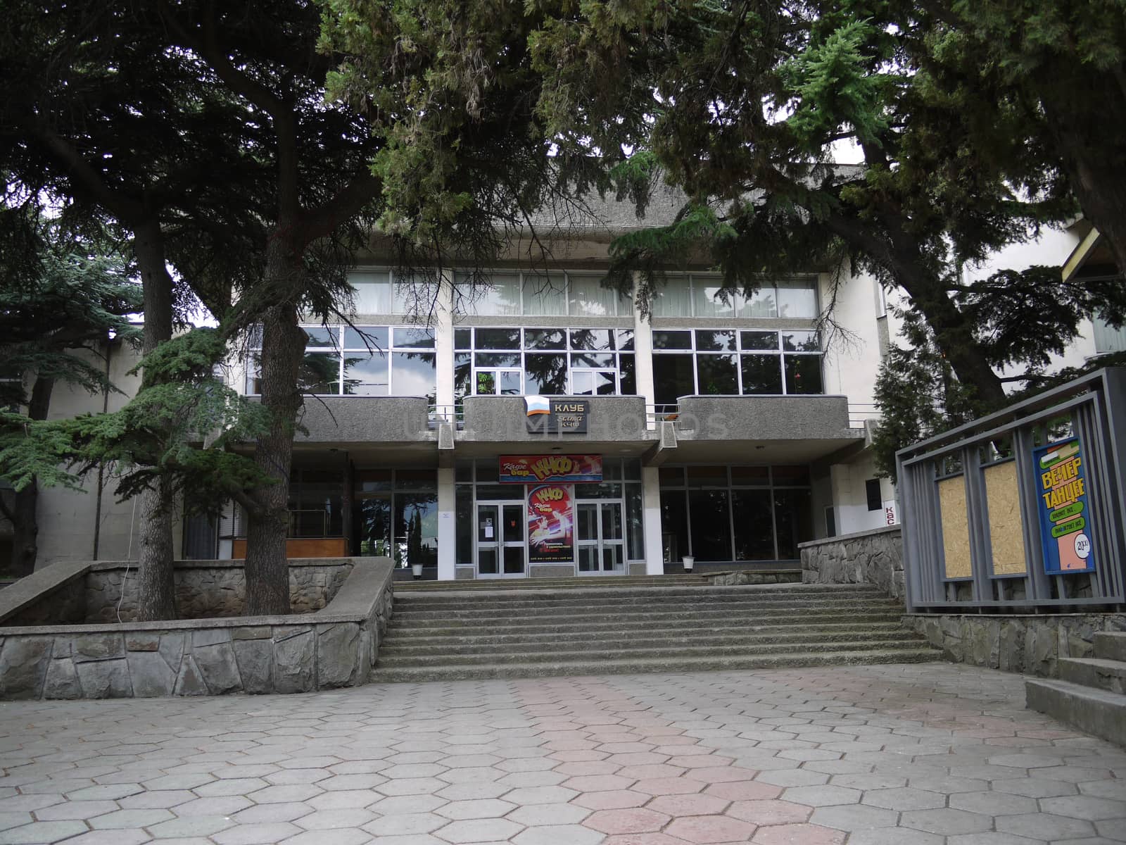 Coniferous trees above the entrance to the two-storeyed house of culture with posters, stairs and a platform in front of the entrance