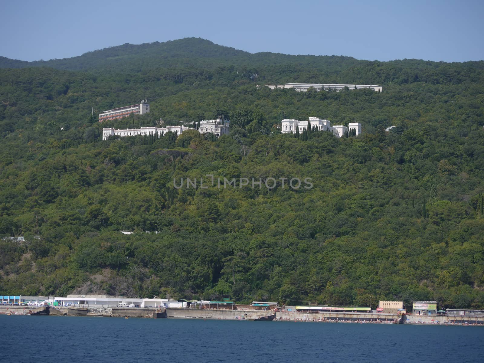 Green mountain slopes covered with dense forest with peeping roofs of houses. The seaside with the beach below. by Adamchuk
