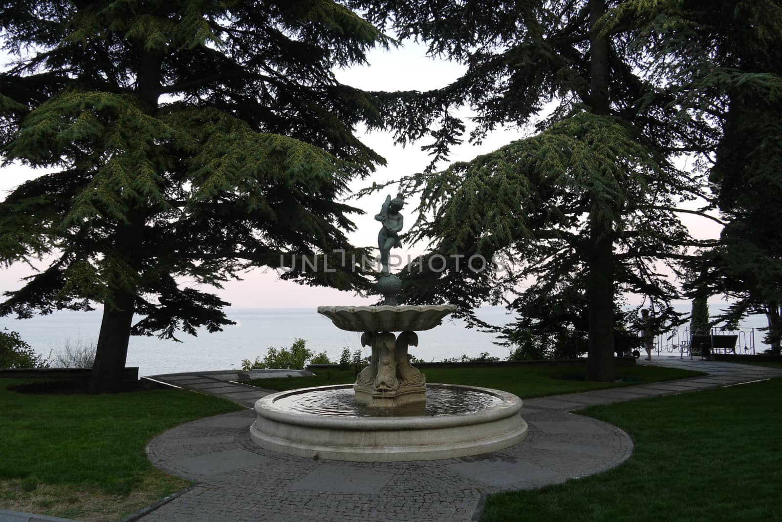 decorative fountain with statue of a girl on the lawn surrounded by fir trees against the background of the sea and sky. place of rest and travel