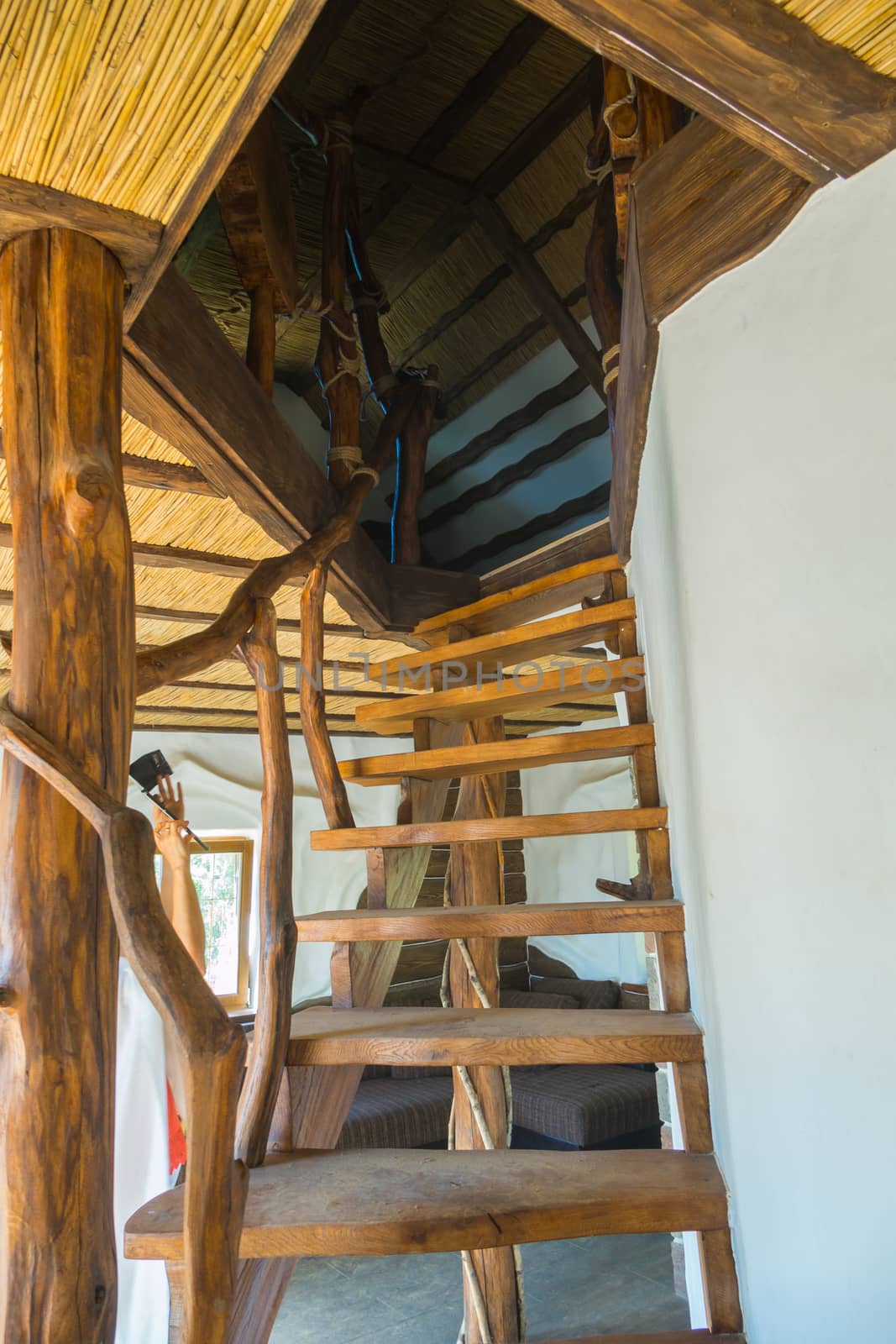 near the white wall stylized decorative unusual wooden staircase with handrails from branches leads to the second floor under a roof covered with straw