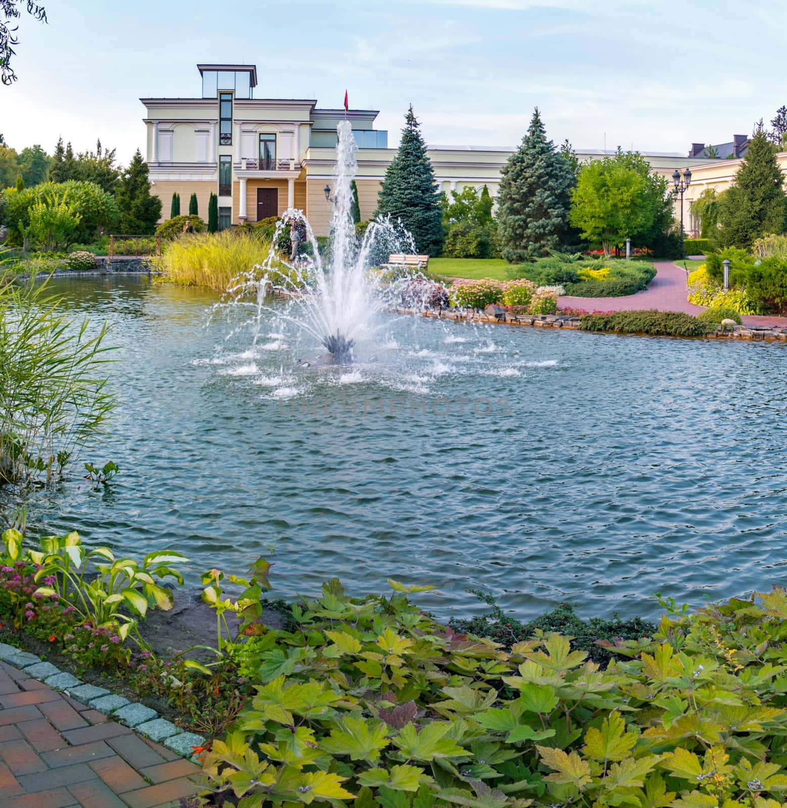 A lovely view of the pond water with a slight ripple on the surface with a fountain in the middle with greenery growing around and a building with a flag standing in the distance.
