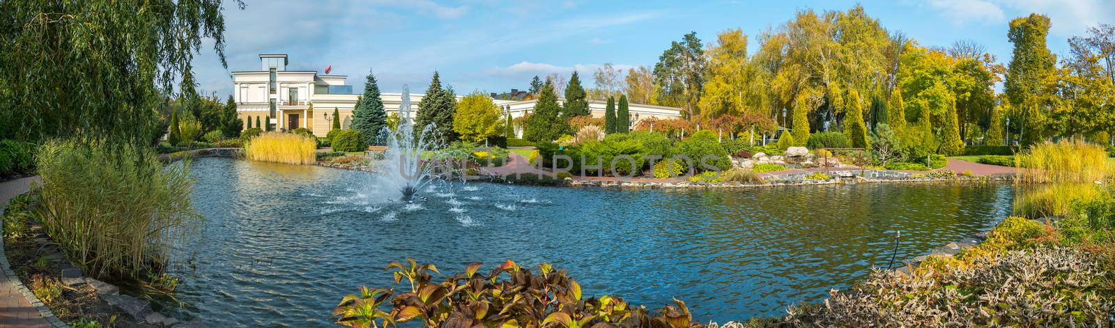 A chic panorama of the park in the summer. With a pond and beating in it a fountain of luxurious green bushes of flowers and trees and a building with a flag fluttering on the roof.