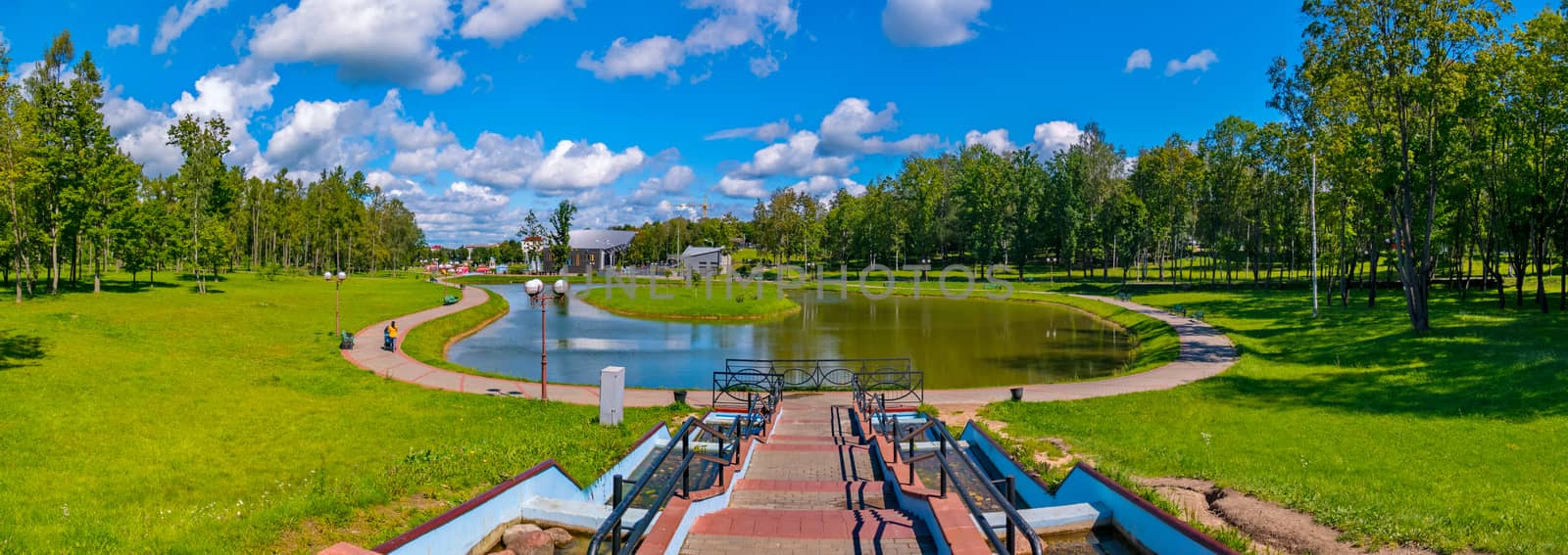 a magnificent panorama of a summer sunny day in a park with a pond and a green island in the middle of it and white clouds floating above this beauty by Adamchuk