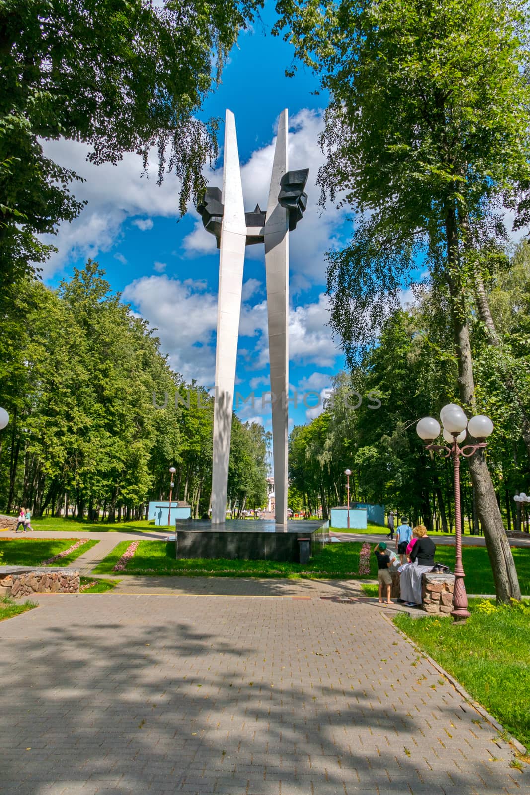 A commemorative sign in a park with resting people in the shade of a tree against a blue sky with slowly floating clouds. by Adamchuk