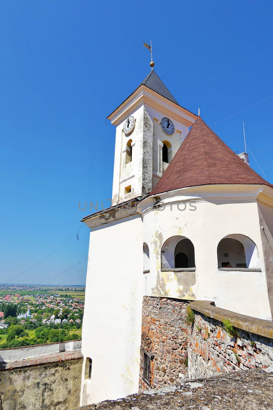 The tower of an ancient Ukrainian castle with a spire and a big clock on a tower in the background of a village with small houses by Adamchuk