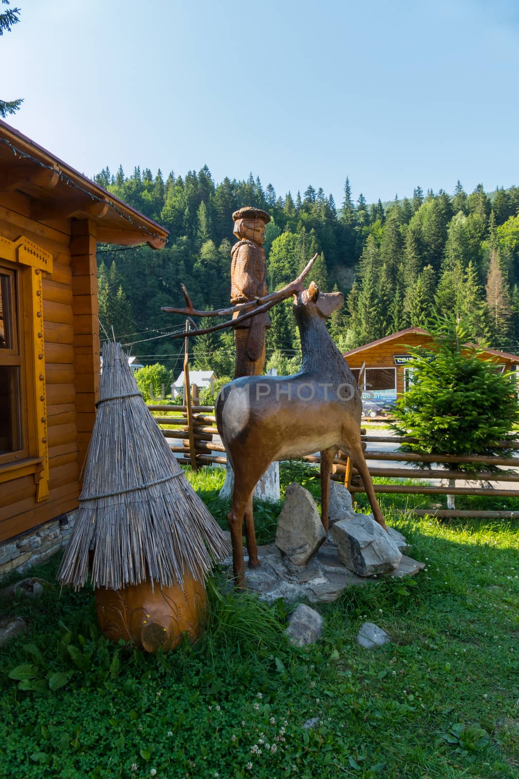 Model of a forest deer on a background of a wooden hut and green coniferous trees