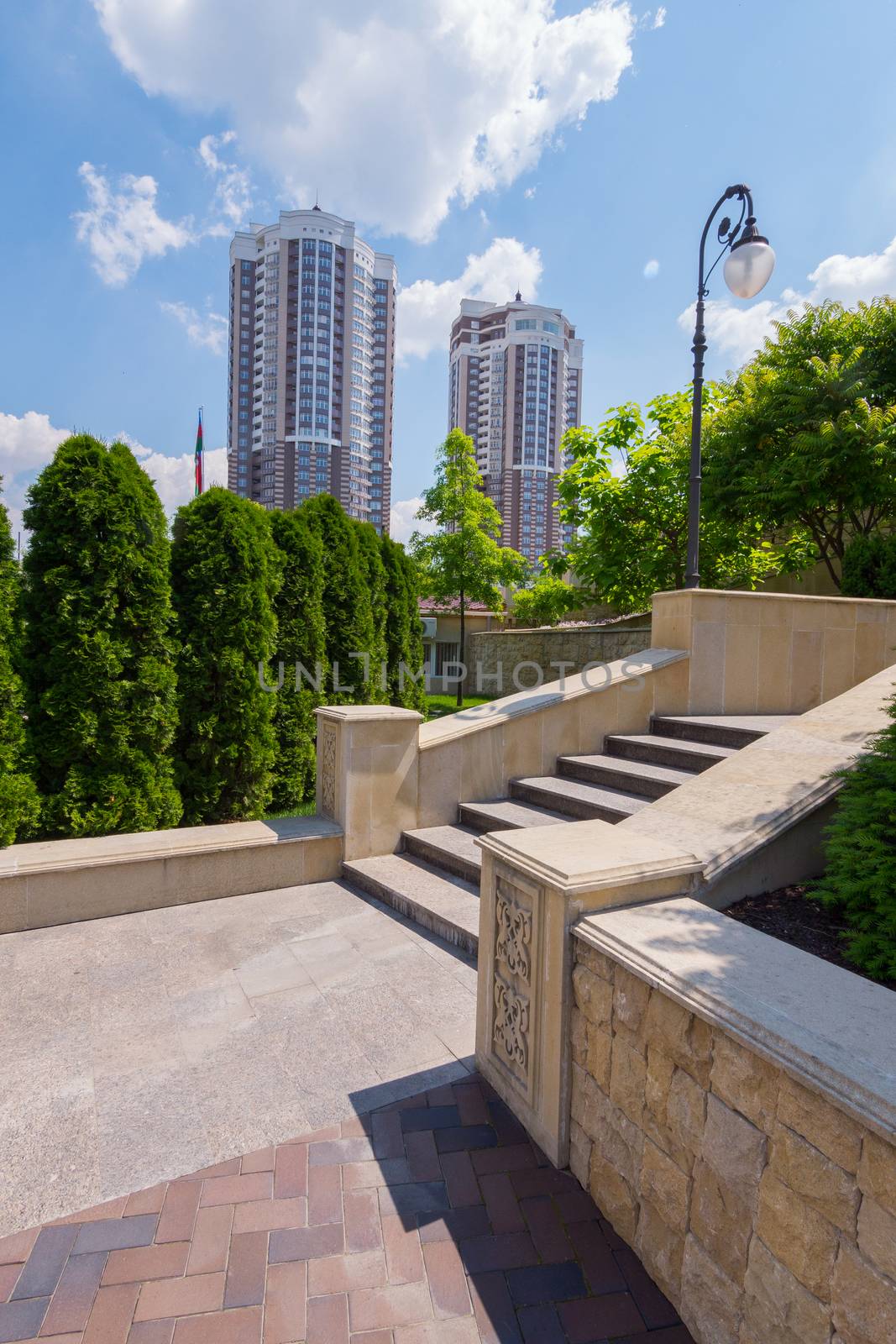 Beautiful view from the platform next to the staircase with steps to two high-rise buildings standing against a background of green trees and a blue sky with clouds. by Adamchuk