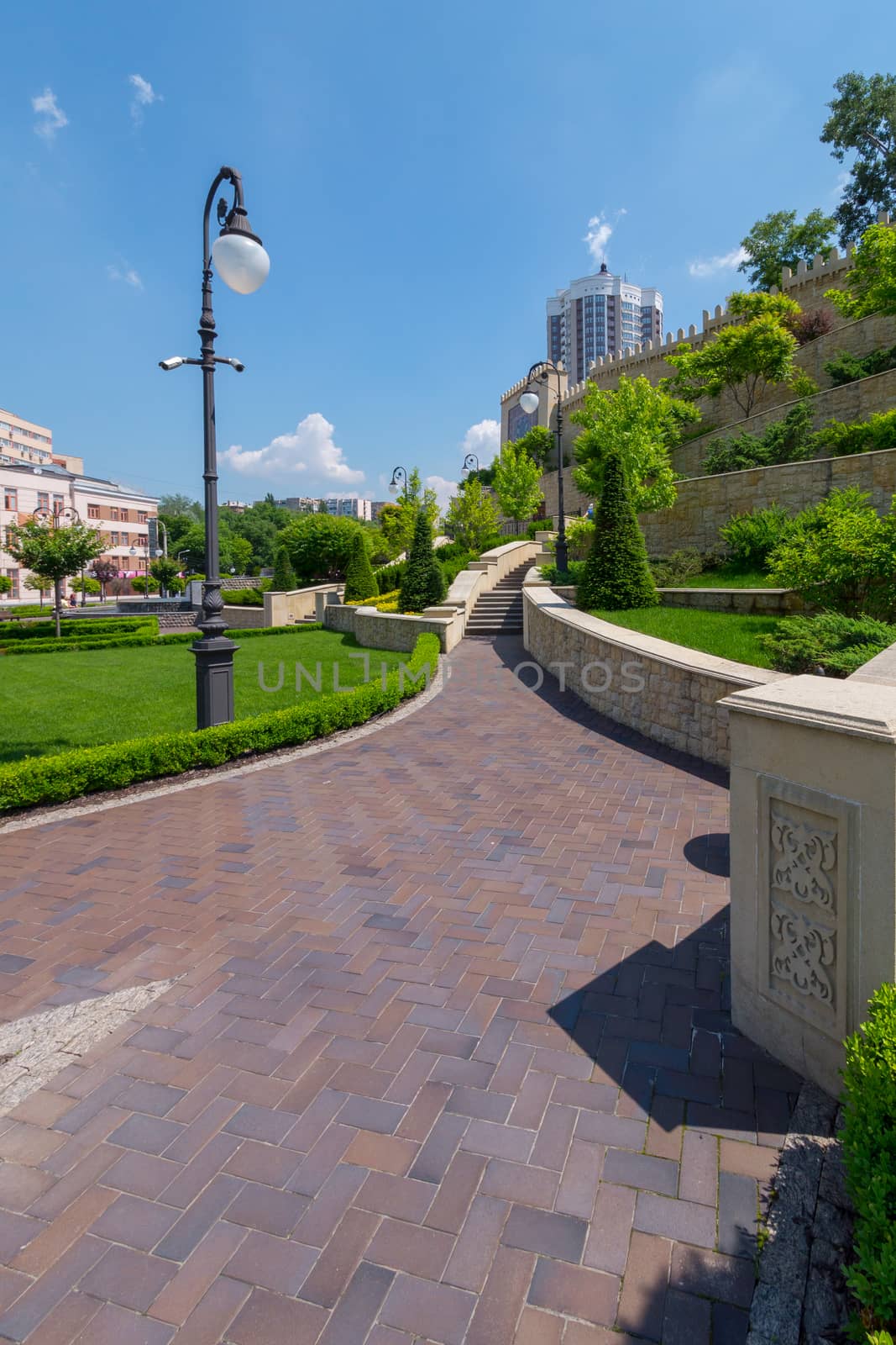 A sunny warm day in a park with a path paved from slabs leading to a multi-level wall with growing trees.