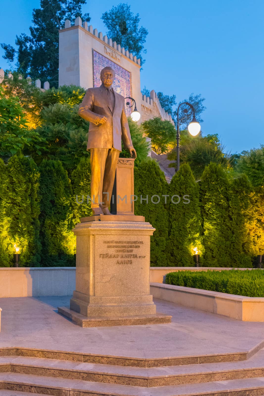 In the evening park with a lighted lantern a monument on the pedestal with a man staring into the distance. Against the background of the wall behind him with patterns and jagged top. by Adamchuk