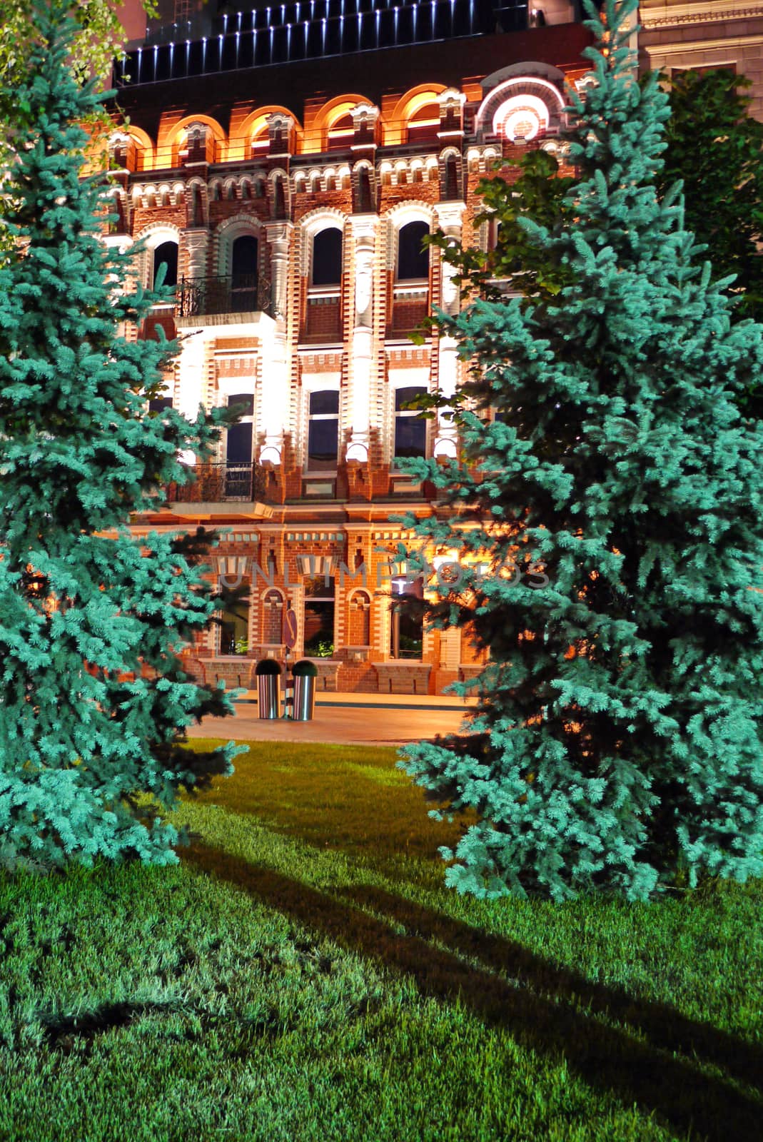 Magnificent view from a green lawn with seated trees on a hotel with beautiful architecture illuminated by evening lights.