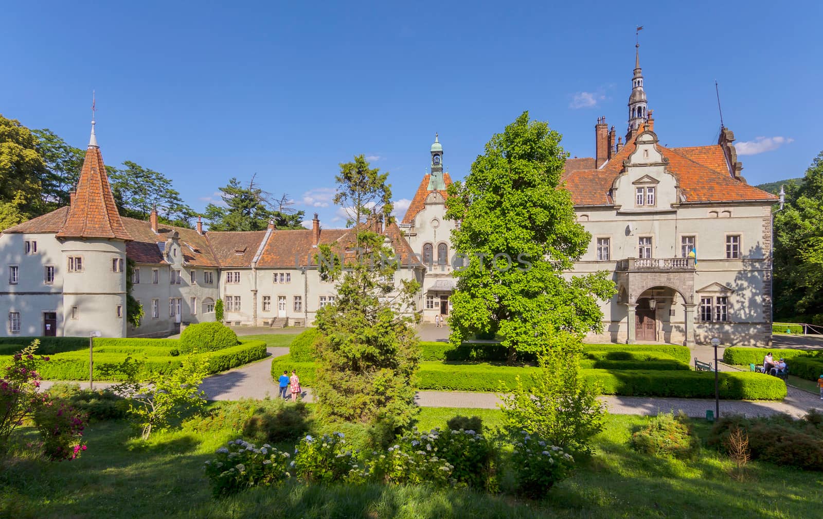 People strolling through the park area near the entrance to the ancient castle with a tiled roof