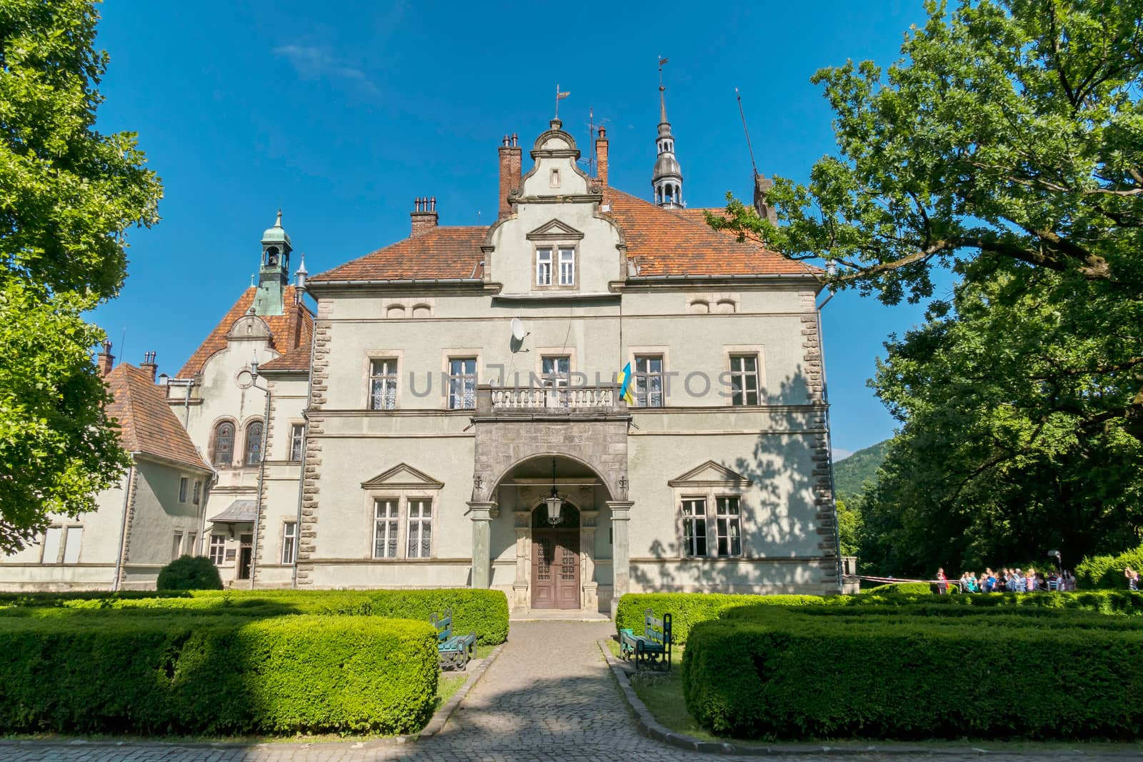 An ancient manor house with a roof from a once red tile. Near it grow bushes and there are two benches opposite each other