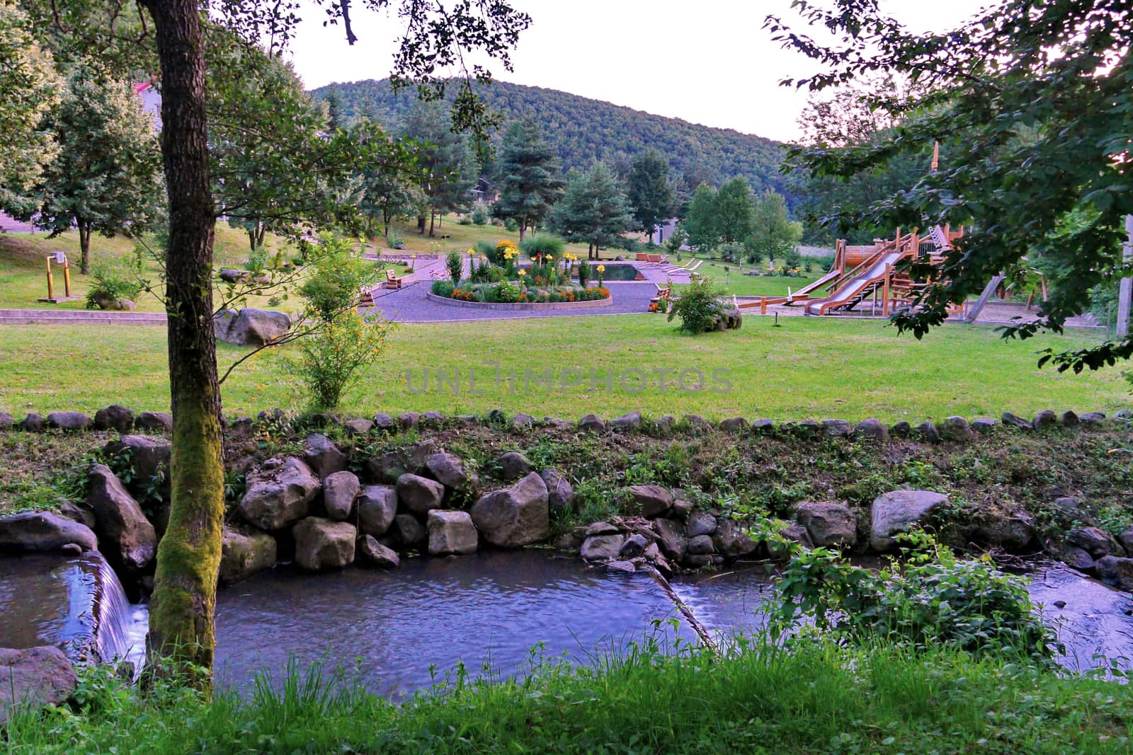 River flow flowing through a small stone canal against the background of a flower bed and a children's playground