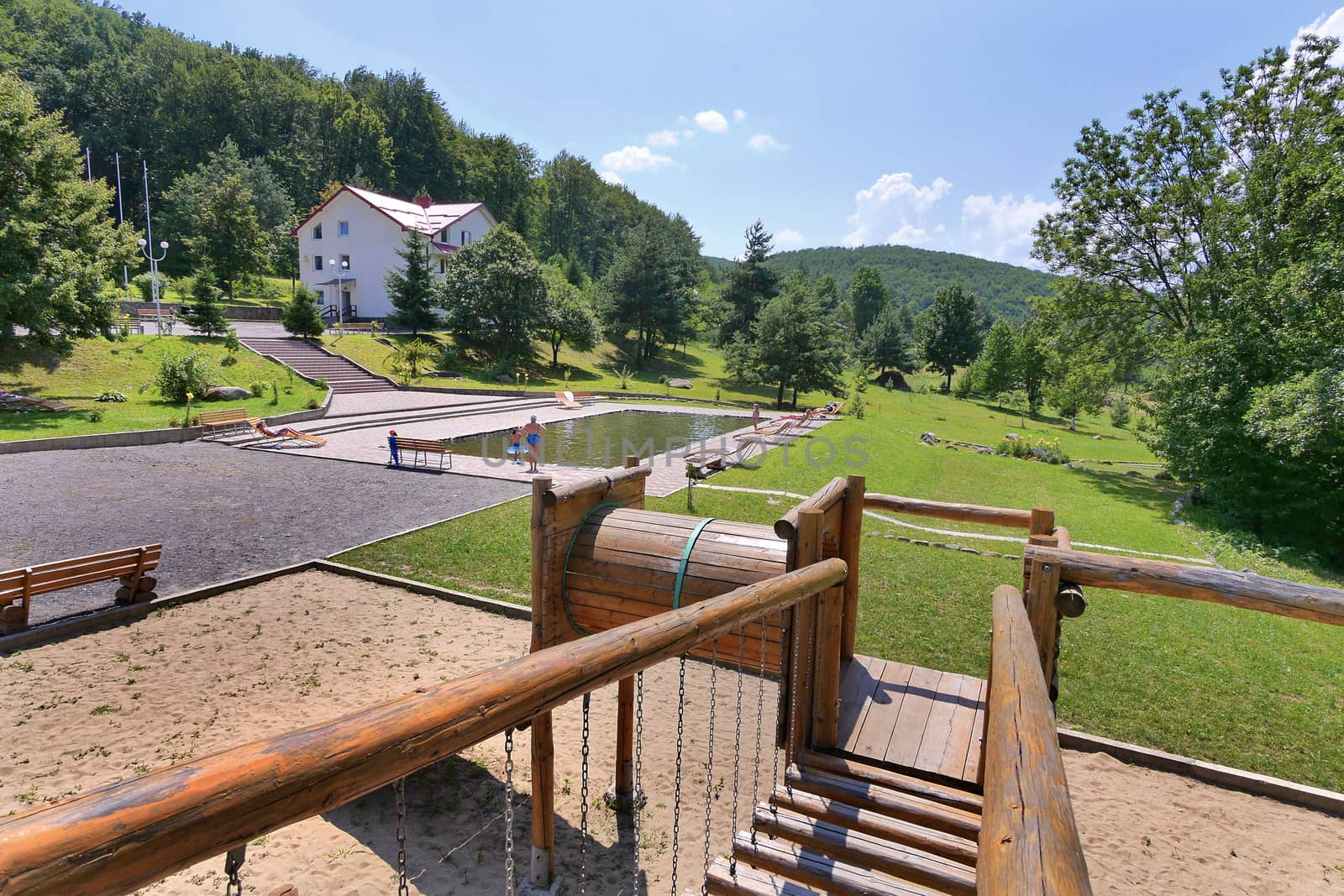 Wooden railing of the dais with a beautiful view of the pool in the park with the resting people with a green lawn nearby and standing in the distance home.