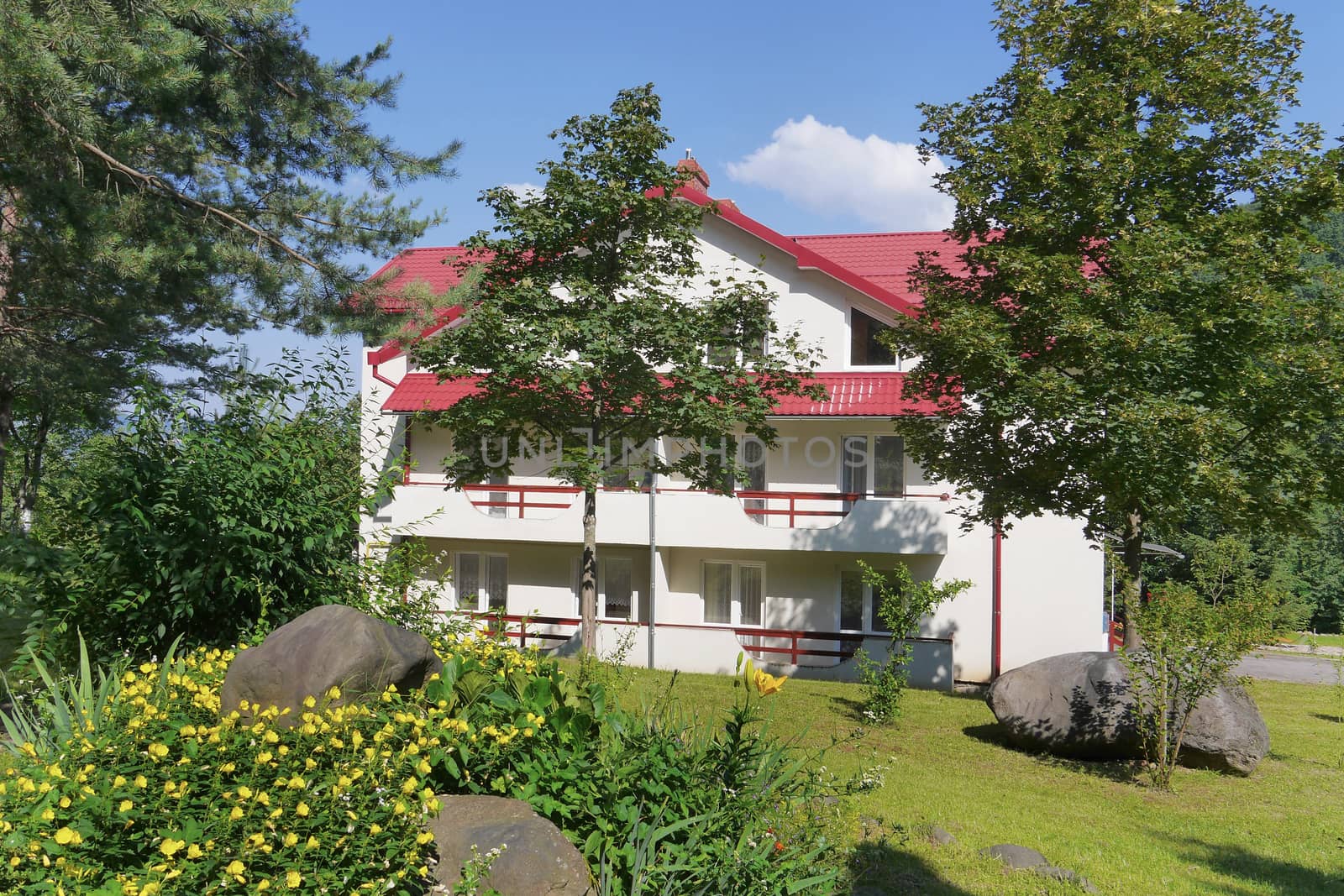 A modern house with white walls and a tiled roof standing amidst the picturesque nature with a green lawn and a designer flower bed with yellow flowers and a stone lying in it.
