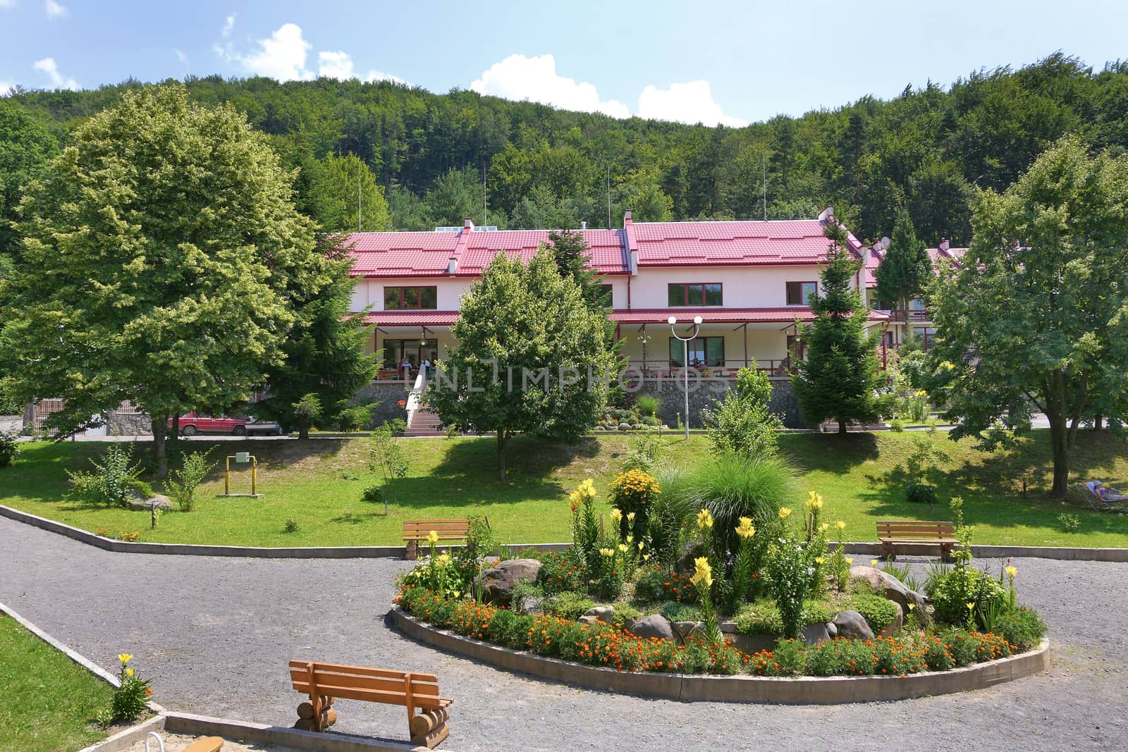A cottage house with a red roof and a park with a circular flower bed in front of it