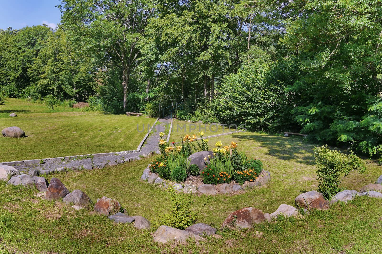 A small bench standing near thick bushes on a green lawn with a beautiful flower bed with flowers surrounded by stones next to it. by Adamchuk