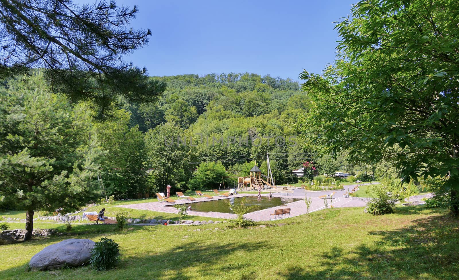 Clean pool with wooden benches, sun loungers and a playground in the background of the park zone