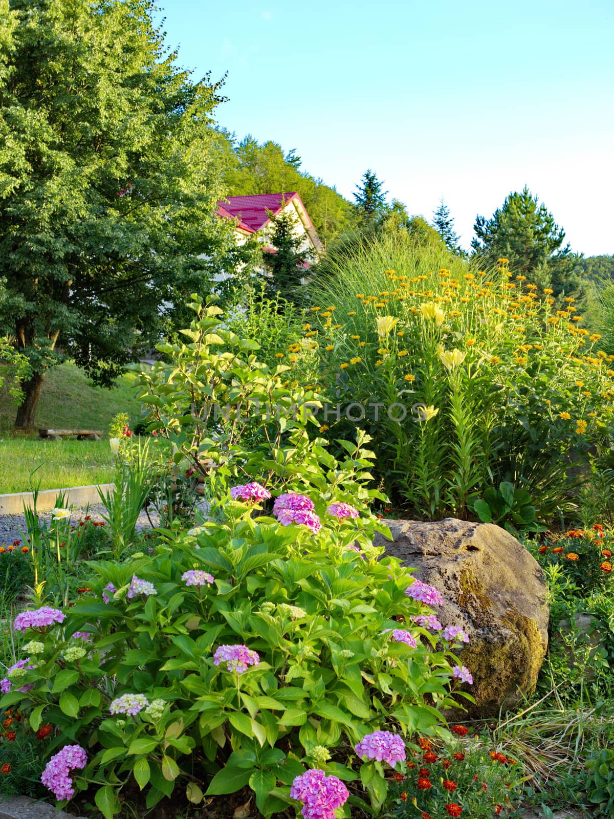 A large stone lying in a flower bed with flowers growing in the open air next to a green lawn and trees. by Adamchuk