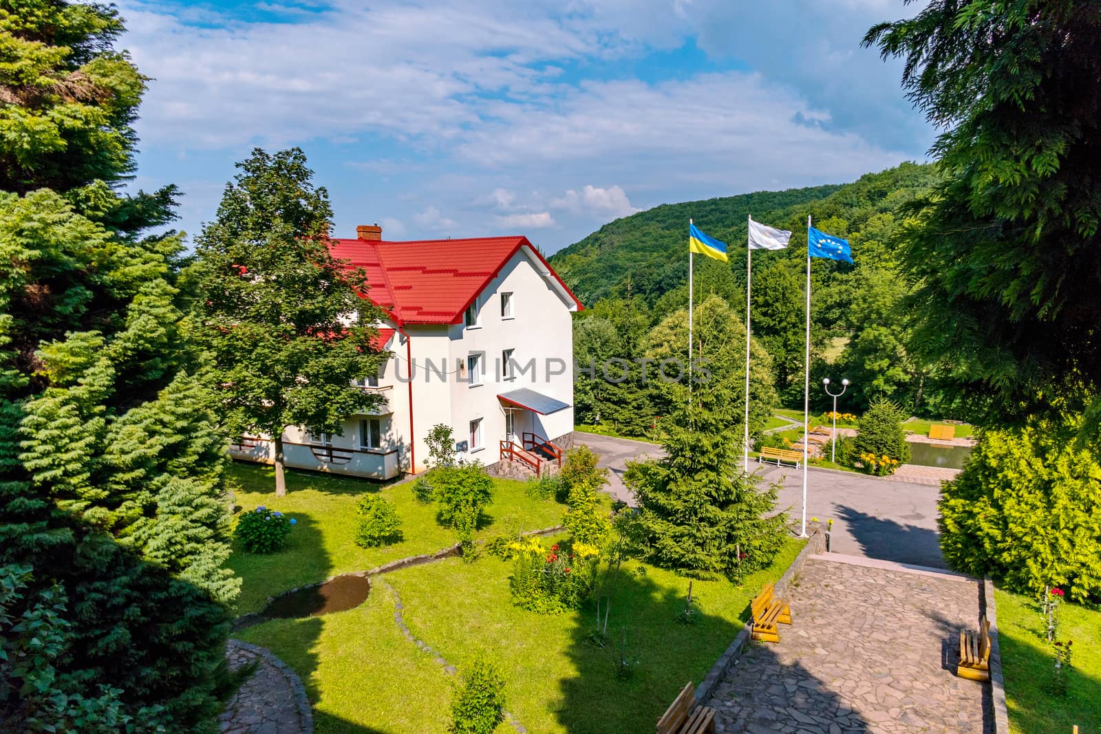 Flags on flagpoles fluttering under the gusts of wind standing next to a modern building against a blue sky with clouds floating above the mountain slopes.