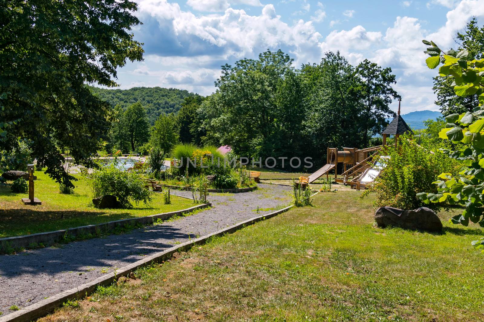 The path leading to the playground near beautiful green flower beds and trees with a swimming pool nearby