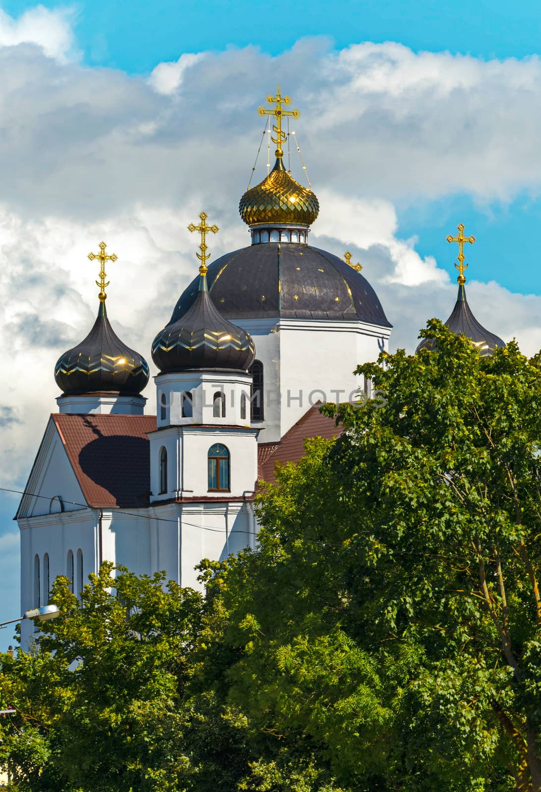 A majestic church with white walls and large domes with golden crosses glistening in the sun, standing among green trees against the sky covered with dense clouds.