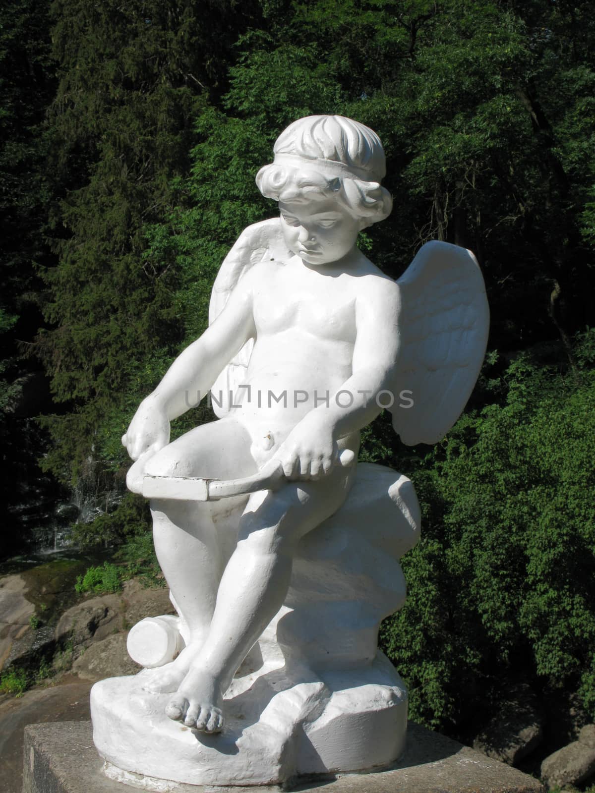 Stone sculpture of Cupid against the background of green deciduous trees