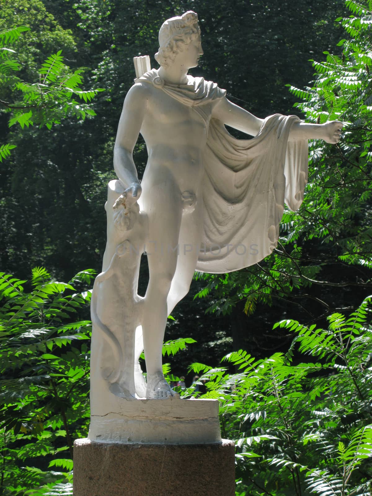 White sculpture of gypsum standing on a pedestal in a park among green bushes illuminated by the rays of a warm summer sun.